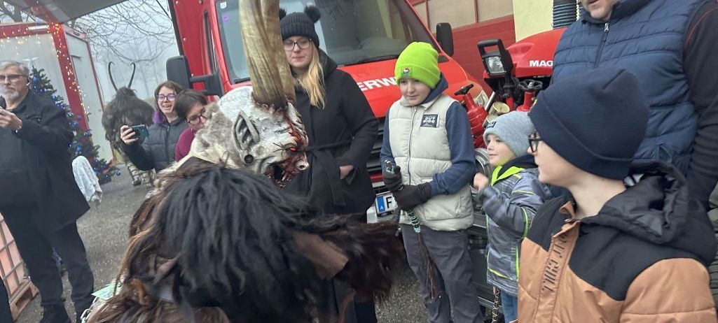 Eine Person in einem gehörnten Kostüm steht vor einem roten Feuerwehrauto. Eine Gruppe von Menschen, darunter Kinder, schaut zu.