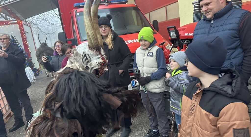 Eine Person in einem gehörnten Kostüm steht vor einem roten Feuerwehrauto. Eine Gruppe von Menschen, darunter Kinder, schaut zu.