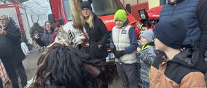 Eine Person in einem gehörnten Kostüm steht vor einem roten Feuerwehrauto. Eine Gruppe von Menschen, darunter Kinder, schaut zu.