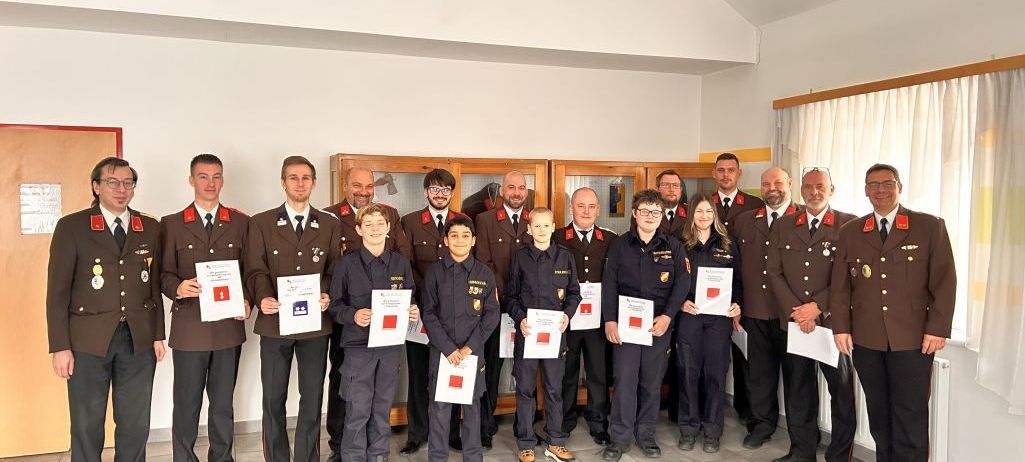 Eine Gruppe von Feuerwehrleuten in Uniform steht zusammen, hält Urkunden in der Hand, einige mit Brille, in einem Raum mit weißen Wänden und gefliesten Böden.