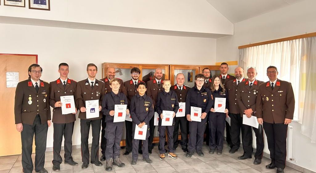 Eine Gruppe von Feuerwehrleuten in Uniform steht zusammen, hält Urkunden in der Hand, einige mit Brille, in einem Raum mit weißen Wänden und gefliesten Böden.