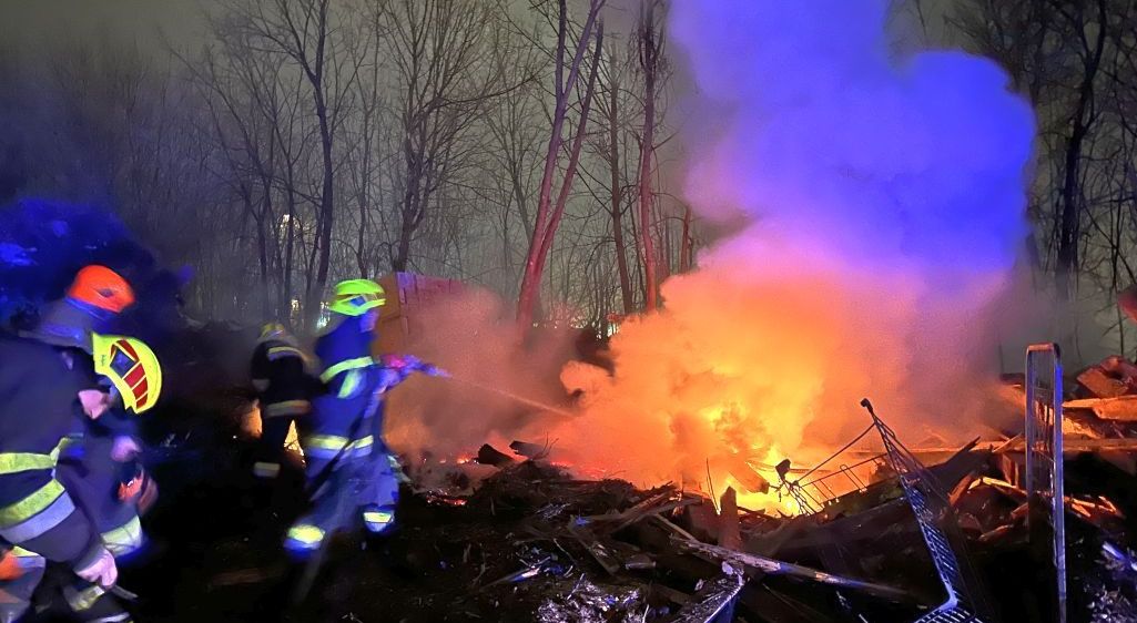 Feuerwehrleute in Schutzkleidung löschen mit einem Schlauch ein Feuer in einem Wald. Bäume und Rauch umgeben die brennenden Trümmer.