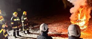 Feuerwehrleute stehen um einen brennenden Weihnachtsbaum herum, wahrscheinlich löschen sie ihn. Die Szene spielt bei Nacht, mit Schnee auf dem Boden.