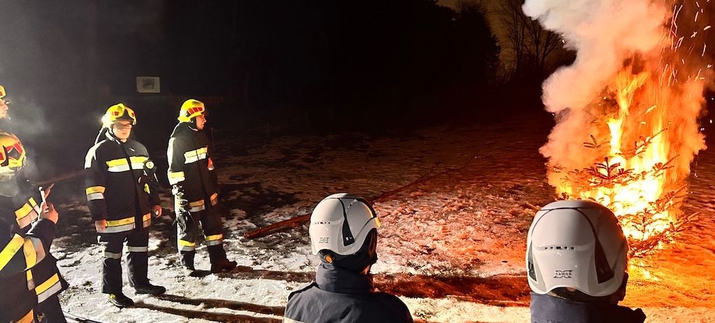 Feuerwehrleute stehen um einen brennenden Weihnachtsbaum herum, wahrscheinlich löschen sie ihn. Die Szene spielt bei Nacht, mit Schnee auf dem Boden.