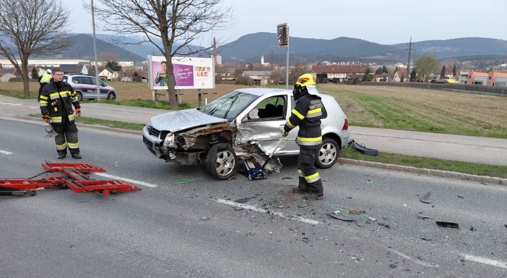 Ein zerkratztes silbernes Auto steht mit Trümmern am Straßenrand. Ein Feuerwehrmann steht daneben, und ein Polizeiauto ist in der Nähe.