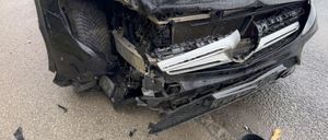 Ein schwarzer Wagen weist starke Frontschäden auf, wobei der Stoßfänger und die Kühlergrill abgerissen sind. Die Motorhaube ist geöffnet, und der Reifen ist verbogen. Trümmerteile sind auf der nassen Straße verstreut.