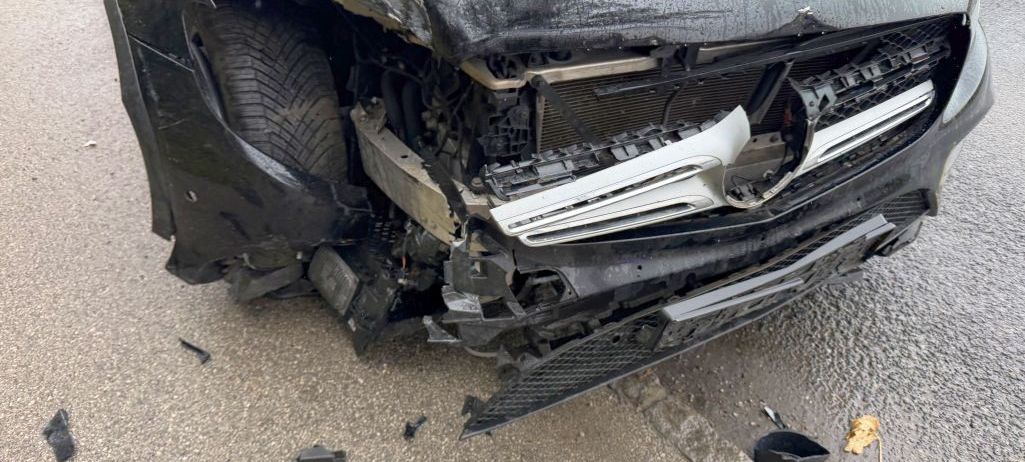 Ein schwarzer Wagen weist starke Frontschäden auf, wobei der Stoßfänger und die Kühlergrill abgerissen sind. Die Motorhaube ist geöffnet, und der Reifen ist verbogen. Trümmerteile sind auf der nassen Straße verstreut.