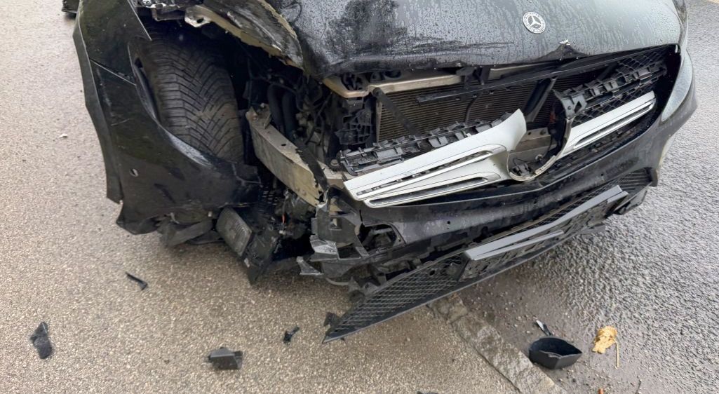 Ein schwarzer Wagen weist starke Frontschäden auf, wobei der Stoßfänger und die Kühlergrill abgerissen sind. Die Motorhaube ist geöffnet, und der Reifen ist verbogen. Trümmerteile sind auf der nassen Straße verstreut.