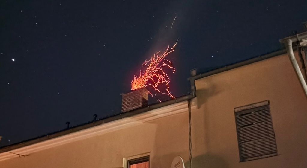 Ein Gebäude mit einem Feuer auf dem Schornstein. Es ist Nacht und der Himmel ist dunkel. Das Gebäude hat Fenster und eine Satellitenschüssel.