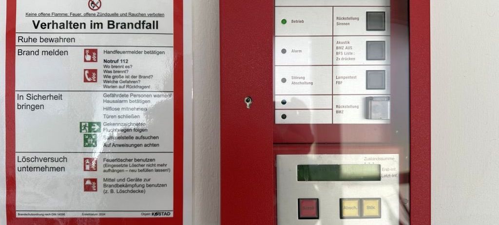 Ein roter Siemens-Feueralarm und ein Feuerschutzhinweis sind an einer Wand angebracht. Der Alarm hat Tasten für Betrieb, Alarm und Rückstellung. Der Hinweis gibt Sicherheitsanweisungen während eines Brandes.