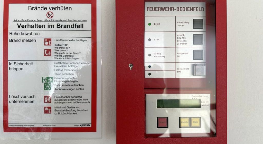 Ein roter Siemens-Feueralarm und ein Feuerschutzhinweis sind an einer Wand angebracht. Der Alarm hat Tasten für Betrieb, Alarm und Rückstellung. Der Hinweis gibt Sicherheitsanweisungen während eines Brandes.