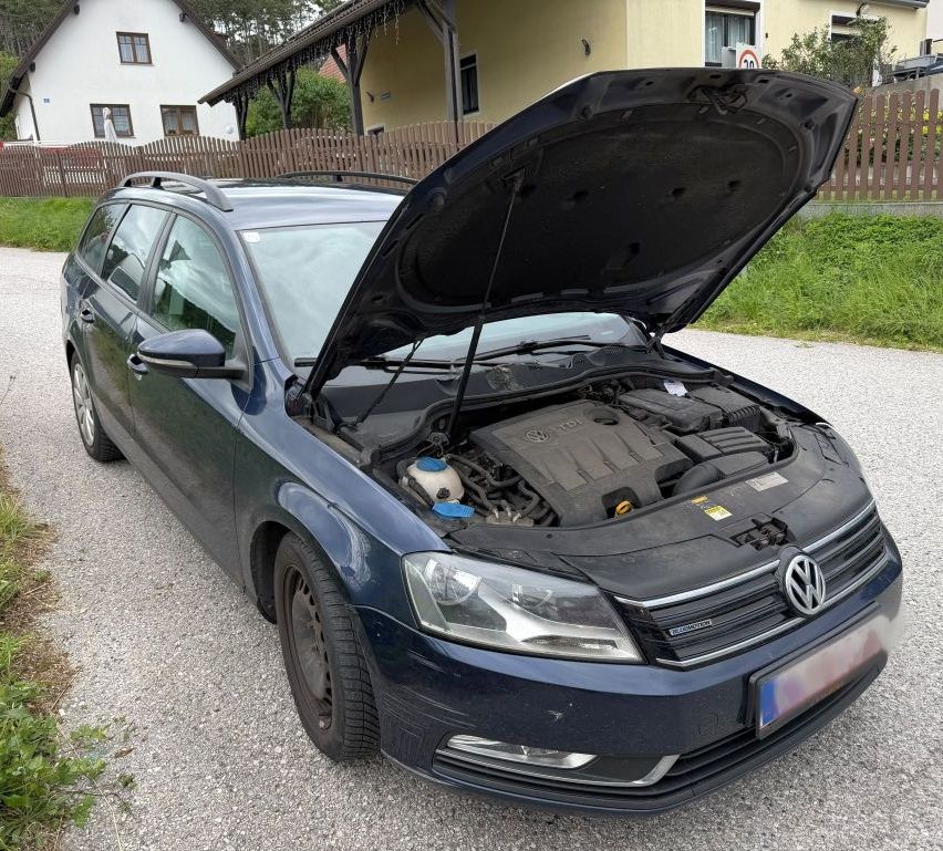A blue Volkswagen Passat with its hood open is parked on gravel near a house with a fence.