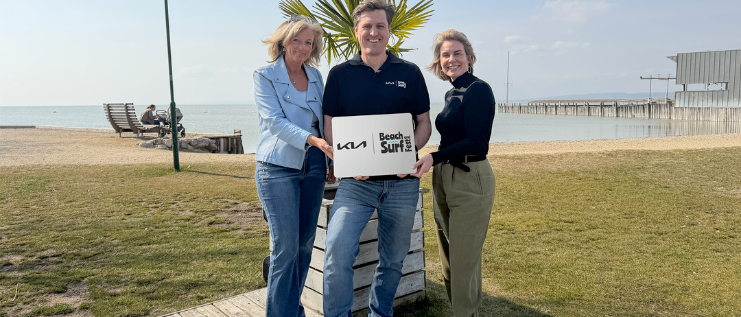 Drei Menschen stehen auf einem Strand, lächeln und halten eine große weiße Box mit der Aufschrift Beach Surf Fest. Dahinter steht eine Palme und ein Holzzaun.