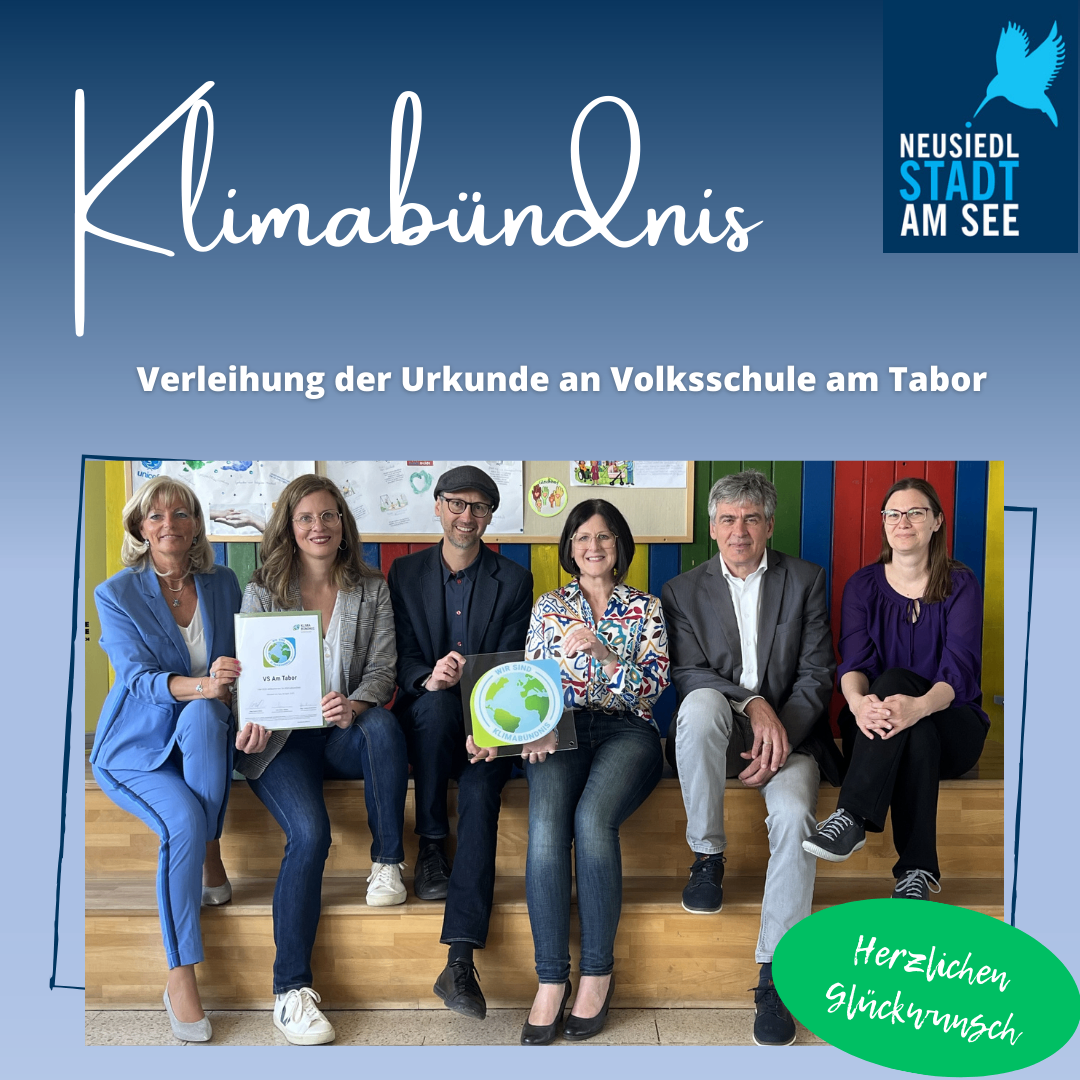 Eine Gruppe Erwachsener posiert mit Urkunden für ein Foto, einer hält einen Globus, vor einer blauen Wand. Der Text lautet: 'Verleihung der Urkunde an Volksschule am Tabor'.