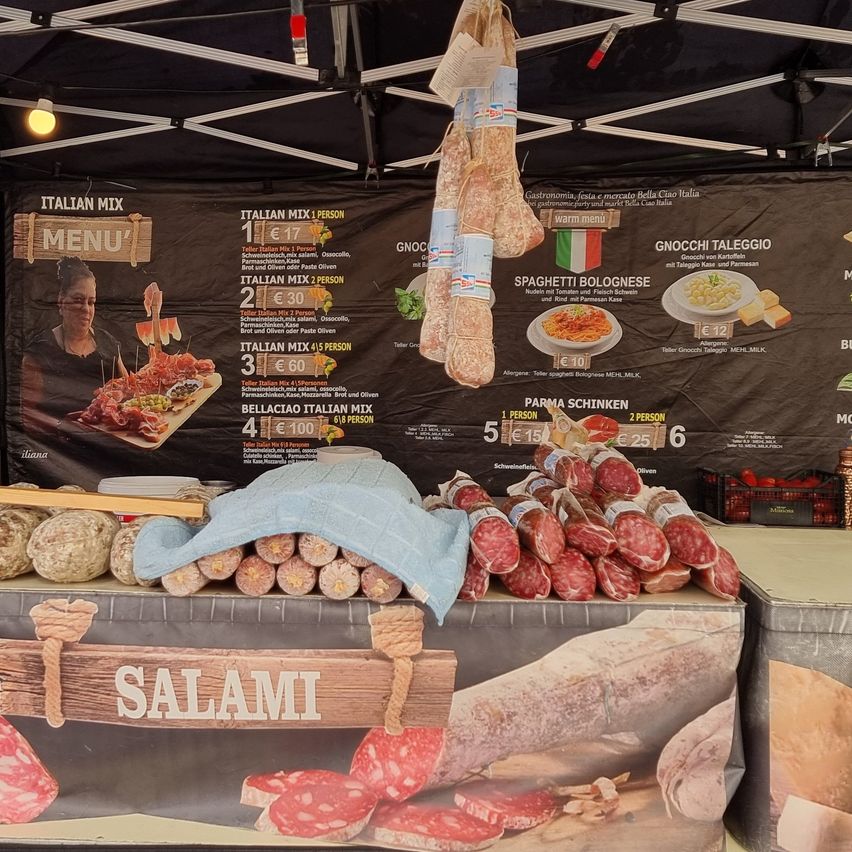 Ein Marktstand im Freien zeigt ein Menü mit italienischen Gerichten, einschließlich Pasta, Fleisch und Käse. Das Menü zeigt Preise für einzelne Portionen und Gruppenbestellungen. Verschiedene Wurstwaren hängen über dem Menü.