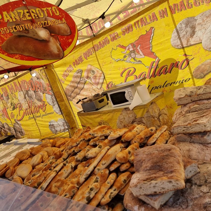 Ein Imbiss präsentiert eine Auswahl an Brot und Gebäck, darunter Sandwiches und Baguettes, unter einem gelben Vordach. Der Stand verfügt über eine Mikrowelle und ein Schild mit der Aufschrift 'Ballando' und einer Karte Italiens.