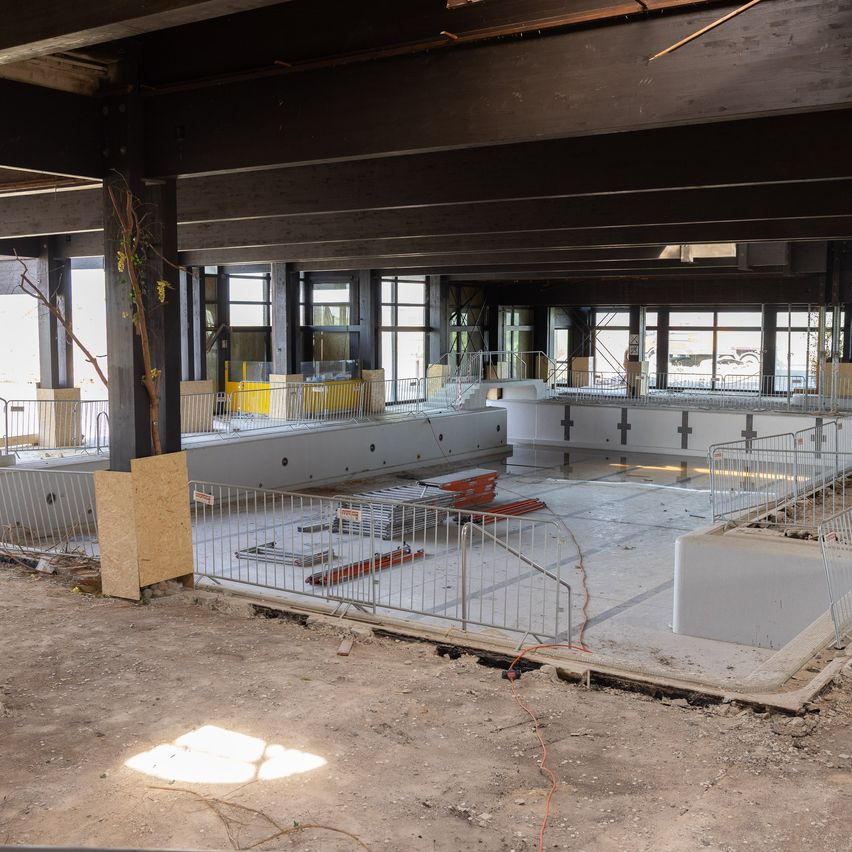 Ein leerer überdachter Pool im Bau mit Metallzäunen und Holzbarrieren. Der Pool ist von einem großen Raum mit mehreren Fenstern umgeben.