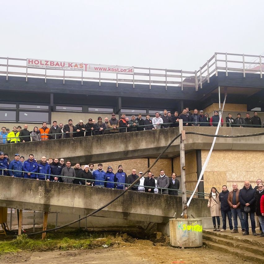 Eine Gruppe von Menschen in Jacken und Mützen steht auf einem Betonbalkon und blickt auf eine Baustelle. Ein Banner mit der Aufschrift 'Holzbau Kast' ist sichtbar. Das Wetter ist bedeckt.