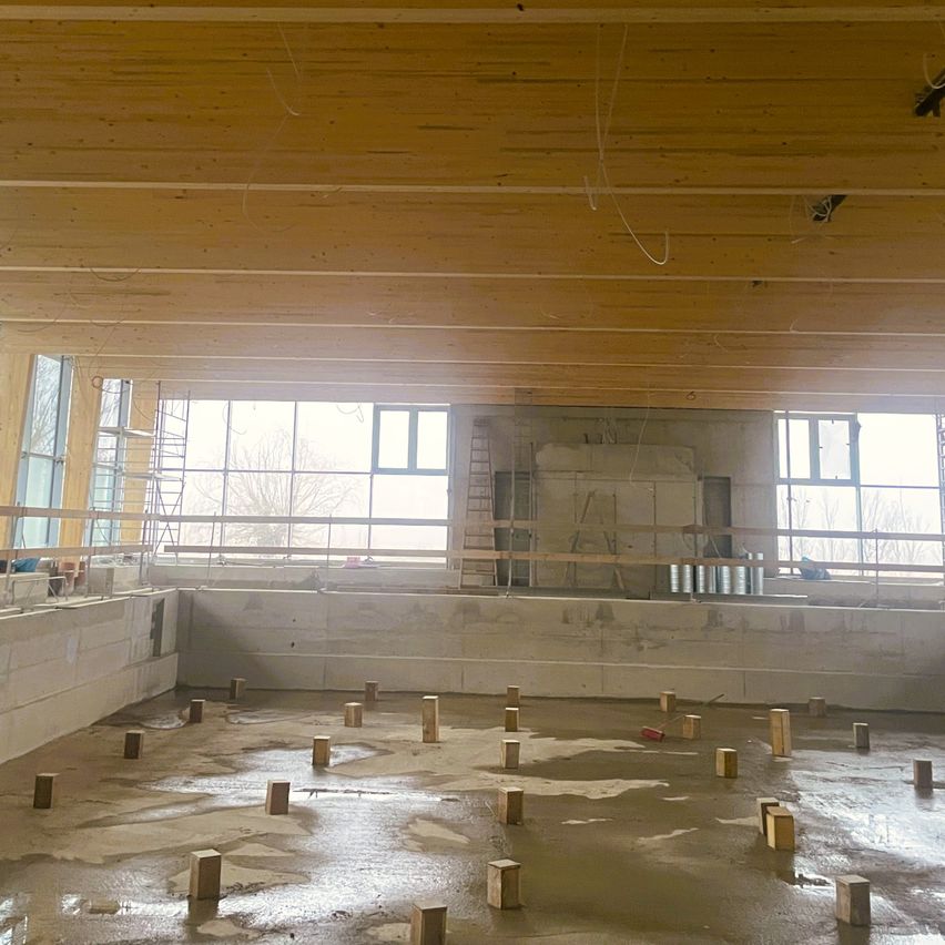 Ein Innenbaustelle mit Holzbalken an der Decke, großen Fenstern und einem nassen Boden. Holzblöcke sind über den Boden verstreut, und eine Leiter lehnt an der Wand.
