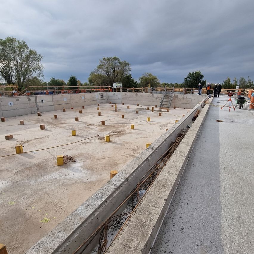 Eine Baustelle mit mehreren Arbeitern in Helmen. Eine große Betonstruktur wird mit gelben Markierungen auf dem Boden gebaut.