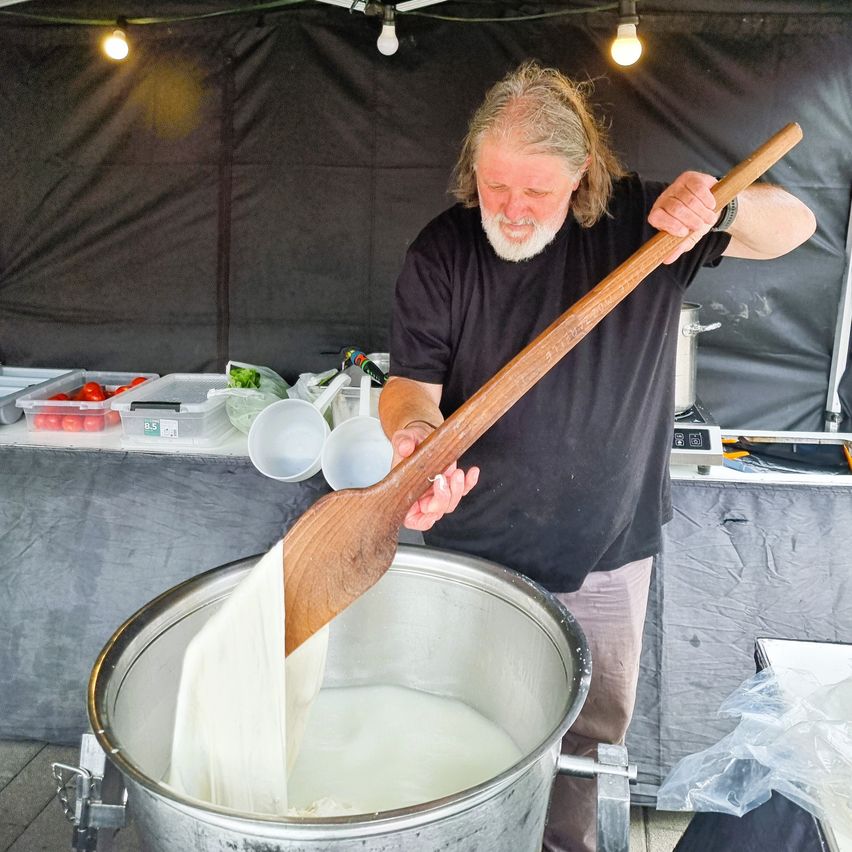 Ein Mann in einem schwarzen T-Shirt rührt mit einem Holzlöffel in einem großen Topf Milch. Er steht in einem Zelt mit einem Tisch dahinter, auf dem verschiedene Lebensmittel stehen.