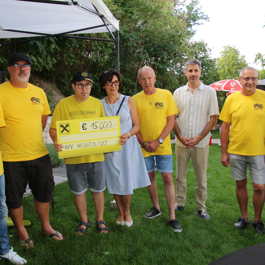 A group of people in yellow shirts stand on grass, one holding a large yellow check. Behind them, a canopy and trees are visible.