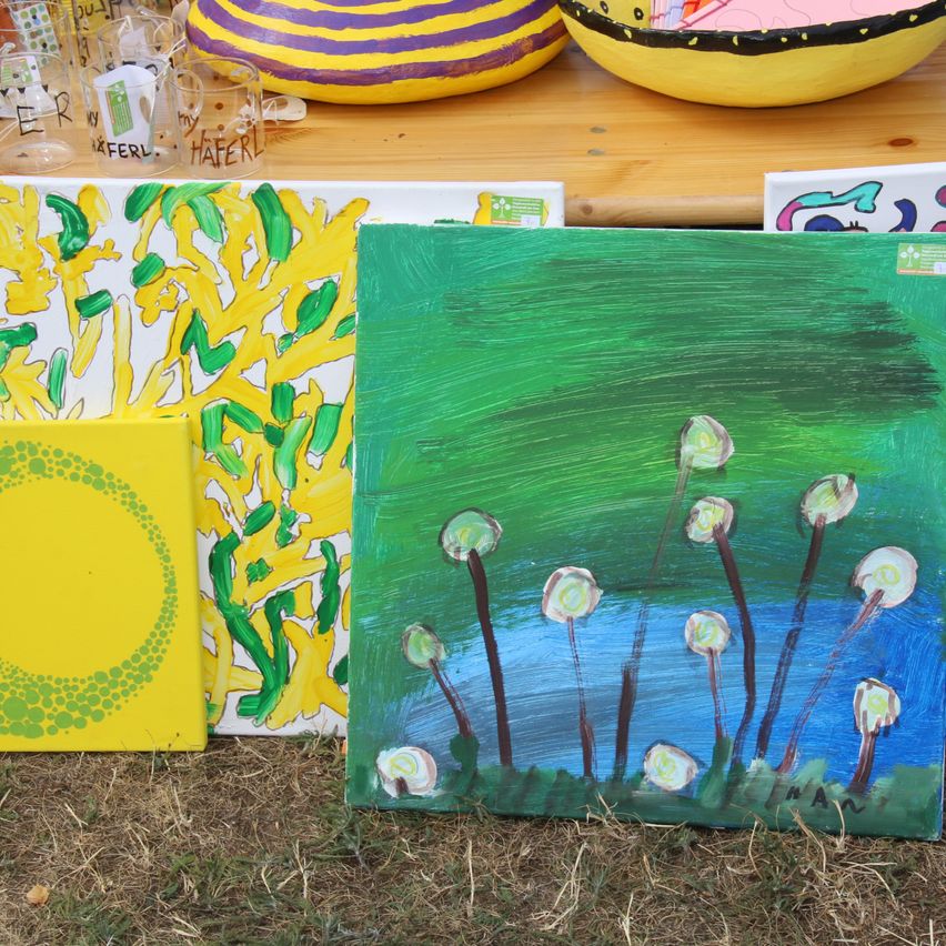 A collection of colorful paintings and ceramic bowls on a wooden table. The paintings feature abstract designs with green, yellow, and blue colors. The ceramic bowls are yellow with black and purple designs.