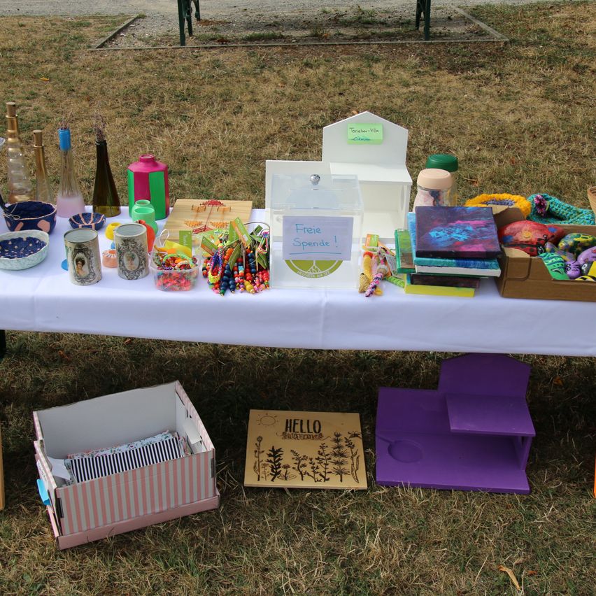 A table covered with a white cloth displays various items including bottles, bowls, and books. A box with the word 'Hello' is on the ground.