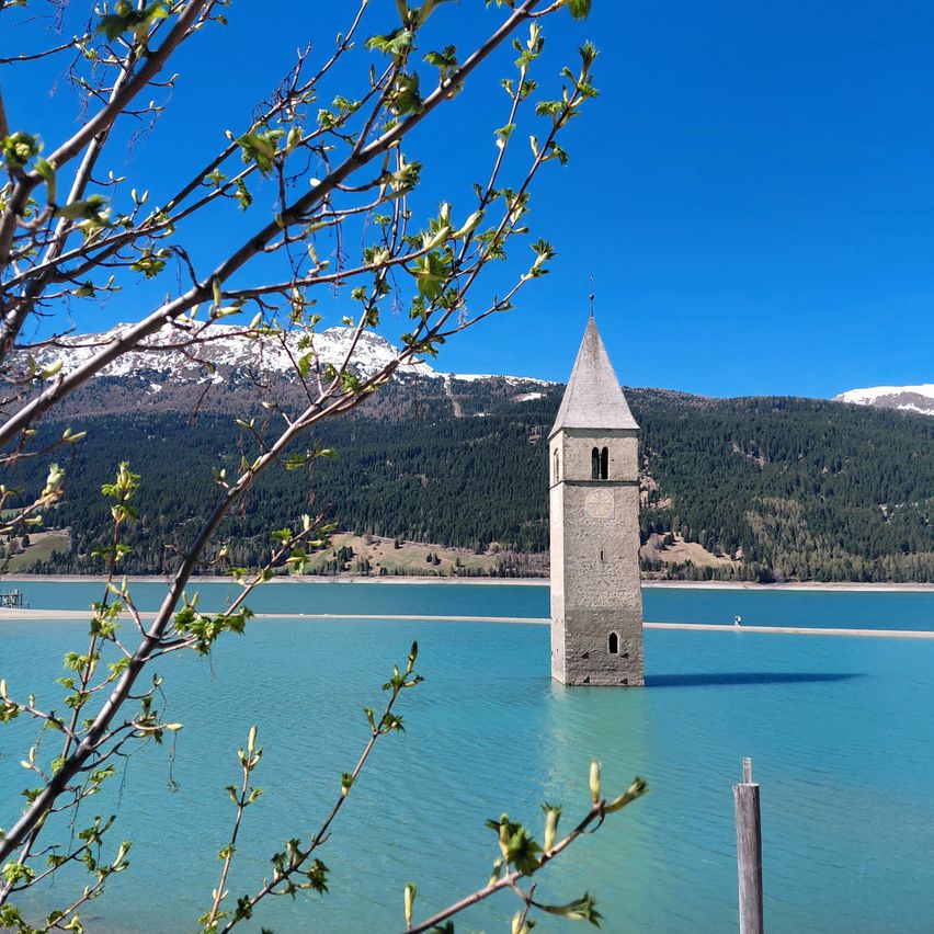 Ein hoher Glockenturm steht allein in der Mitte eines Sees mit Bergen im Hintergrund.