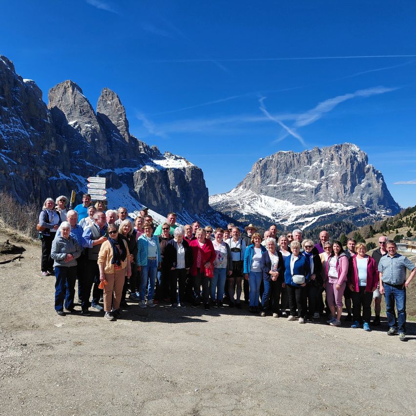Eine große Gruppe von Wanderern steht vor einer Bergkulisse, alle mit unterschiedlicher Outdoor-Ausrüstung. Der Himmel ist klar und blau mit verstreuten Wolken. Die Gruppe posiert für ein Foto.