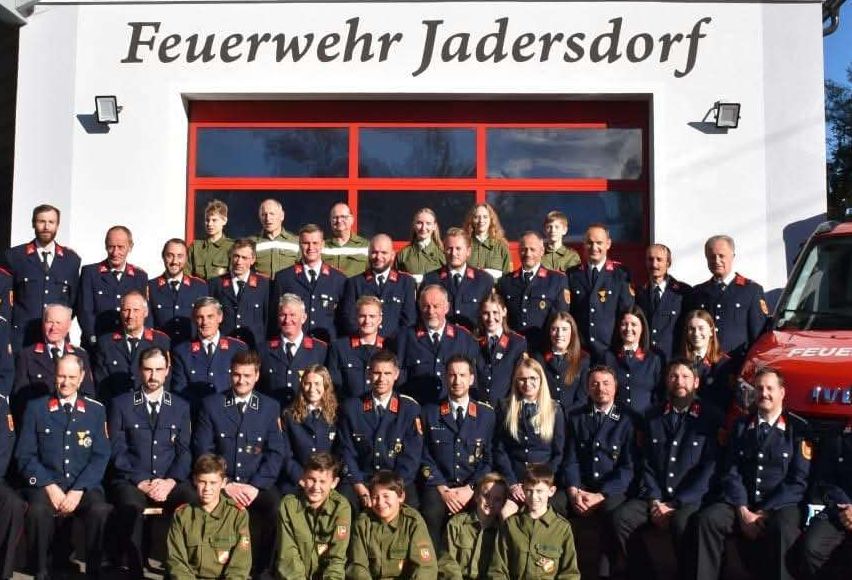 Eine Gruppe von Feuerwehrleuten der Feuerwehr Jadersdorf posiert vor ihrem Feuerwehrhaus.