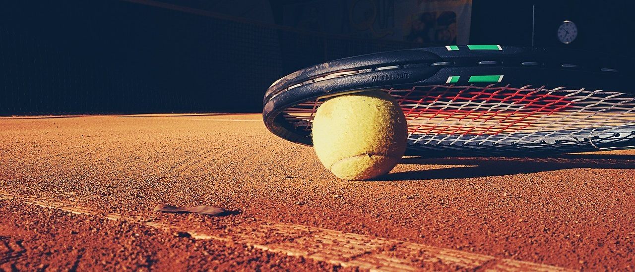 Ein Tennisball liegt auf einem Ascheplatz, mit einem Tennisschläger in der Nähe, das einen Schatten auf den Platz wirft.