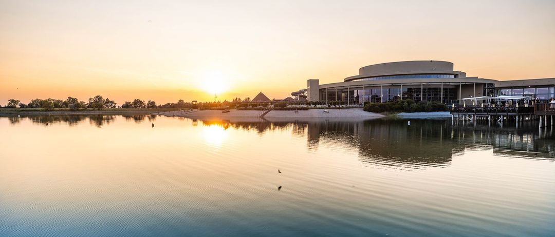 Sonnenuntergang über einem ruhigen See mit einem modernen Gebäude und einer kleinen Pyramidenstruktur am Ufer.