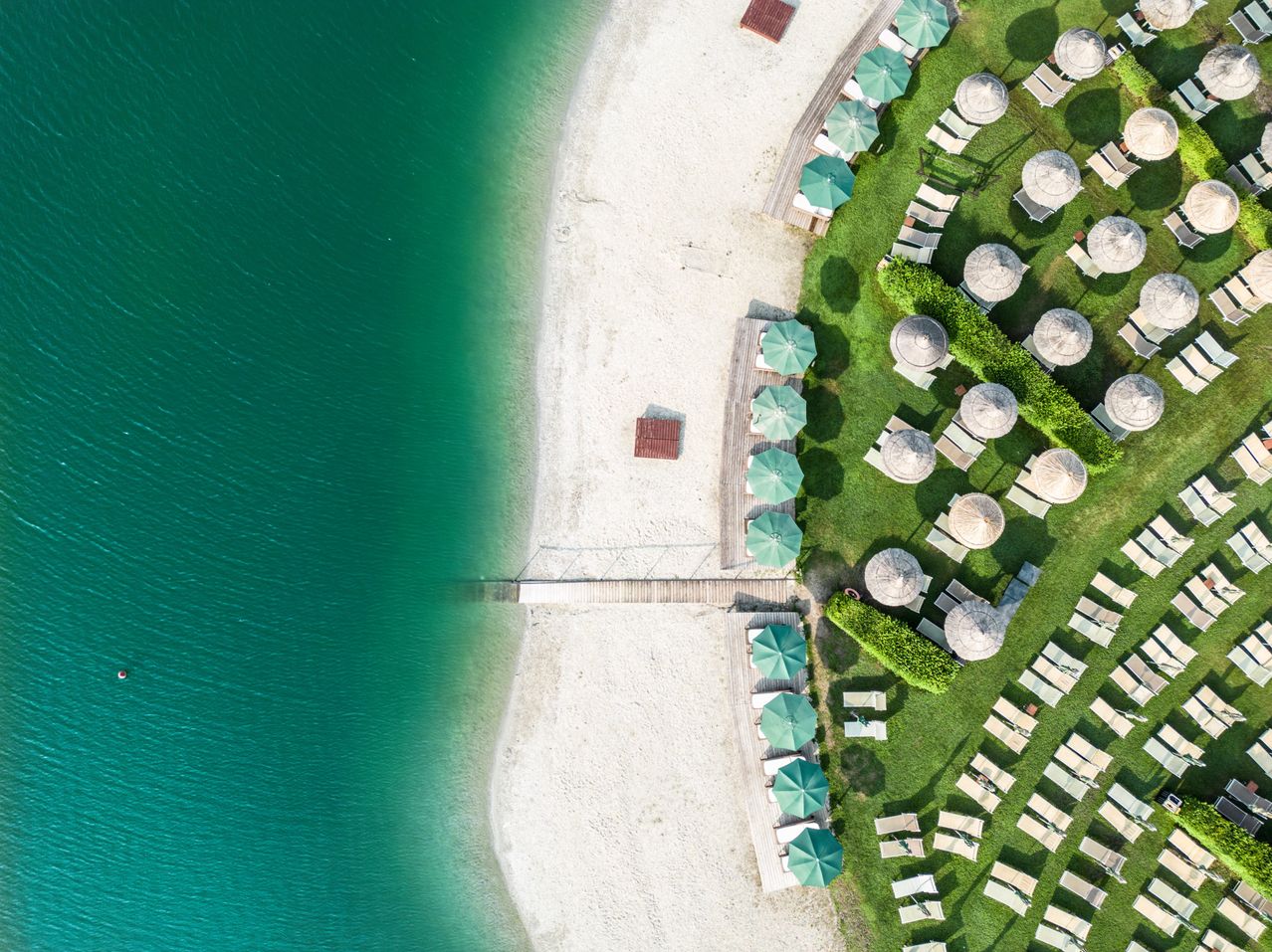 Luftaufnahme eines Strandes mit weißem Sand, grünem Gras und Sonnenschirmen am Ufer.