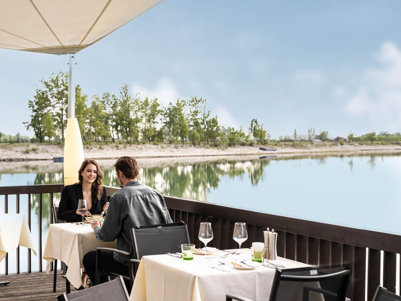 Ein Paar sitzt auf einer Terrasse eines Restaurants in der Nähe eines Sees und genießt eine Mahlzeit mit Weingläsern.