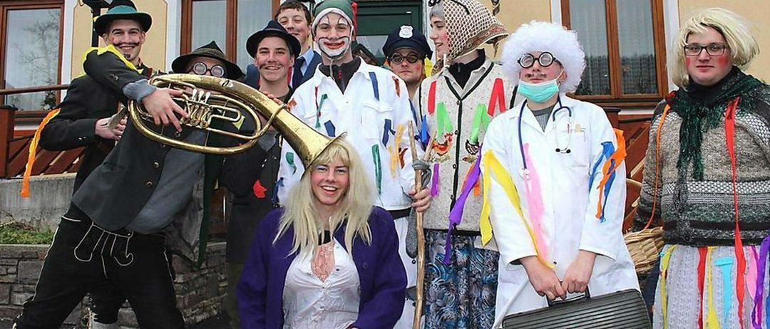 Eine Gruppe von Menschen in verschiedenen Kostümen posiert für ein Foto vor einem Gebäude. Das Schild darüber lautet Gasthof Purrer. Die Kostüme umfassen einen Clown, einen Arzt und andere.