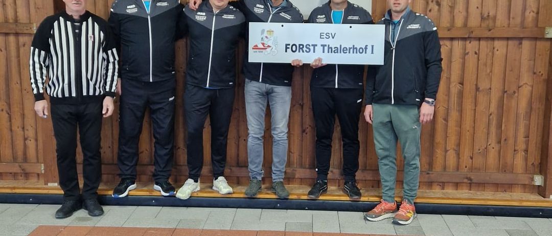 Fünf Männer stehen vor einer Holzwand. Sie tragen schwarze Jacken und einer trägt ein gestreiftes Hemd. Sie halten ein Schild mit der Aufschrift Forst Thalerhof I.