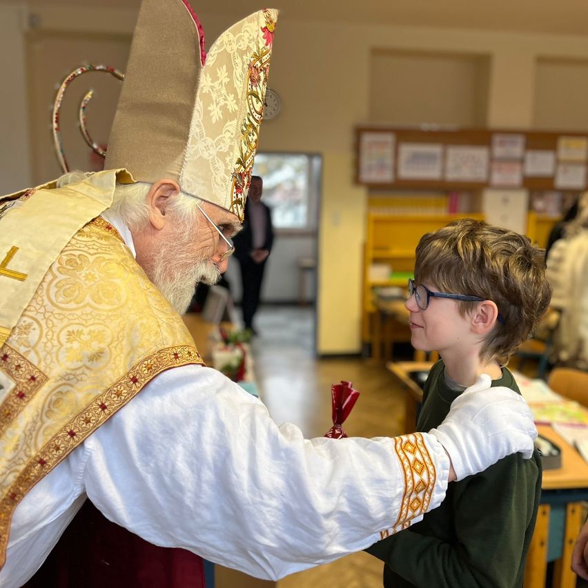 Ein älterer Mann in geistlicher Kleidung gibt einem jungen Jungen in einem Klassenzimmer ein rotes Band. Der Junge trägt eine Brille und ein grünes Hemd. Der Raum hat Schreibtische und Anschlagtafeln.