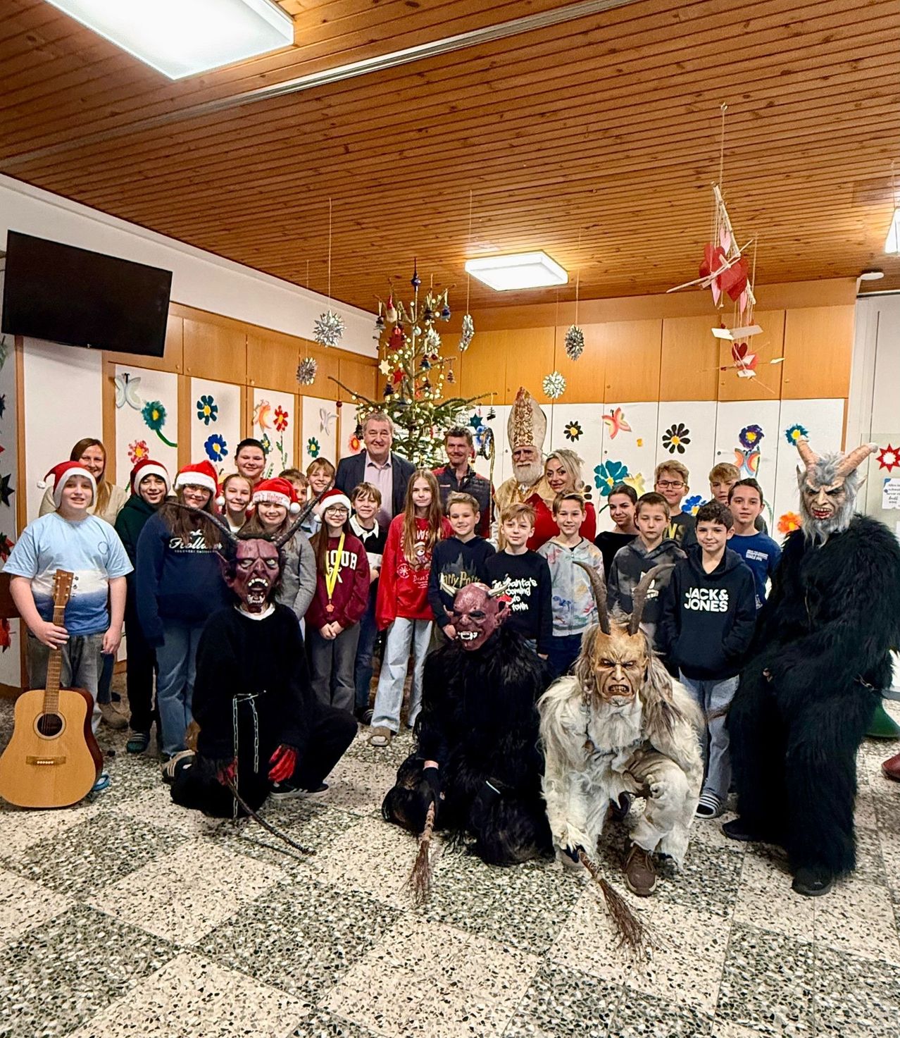 Eine Gruppe von Menschen posiert für ein Foto. Einige tragen Weihnachtsmützen. Zwei Personen in Kostümen scheinen gruselig zu sein. Ein Weihnachtsbaum steht hinter ihnen. Einige Leute halten Gitarren. Ein Monitor ist an der Wand montiert.