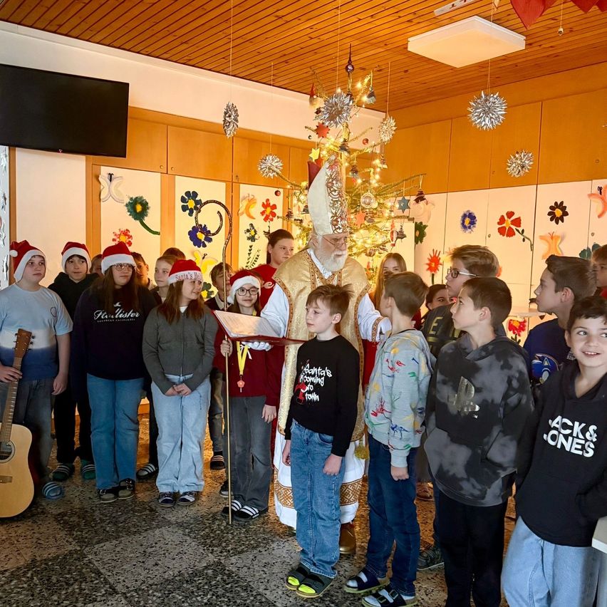 Ein Priester in traditioneller Kleidung liest aus einem Buch, während eine Gruppe von Kindern in Winterkleidung aufmerksam zuhört. Der Raum ist mit einem Baum, Blumen und hängenden Dekorationen für Weihnachten geschmückt.