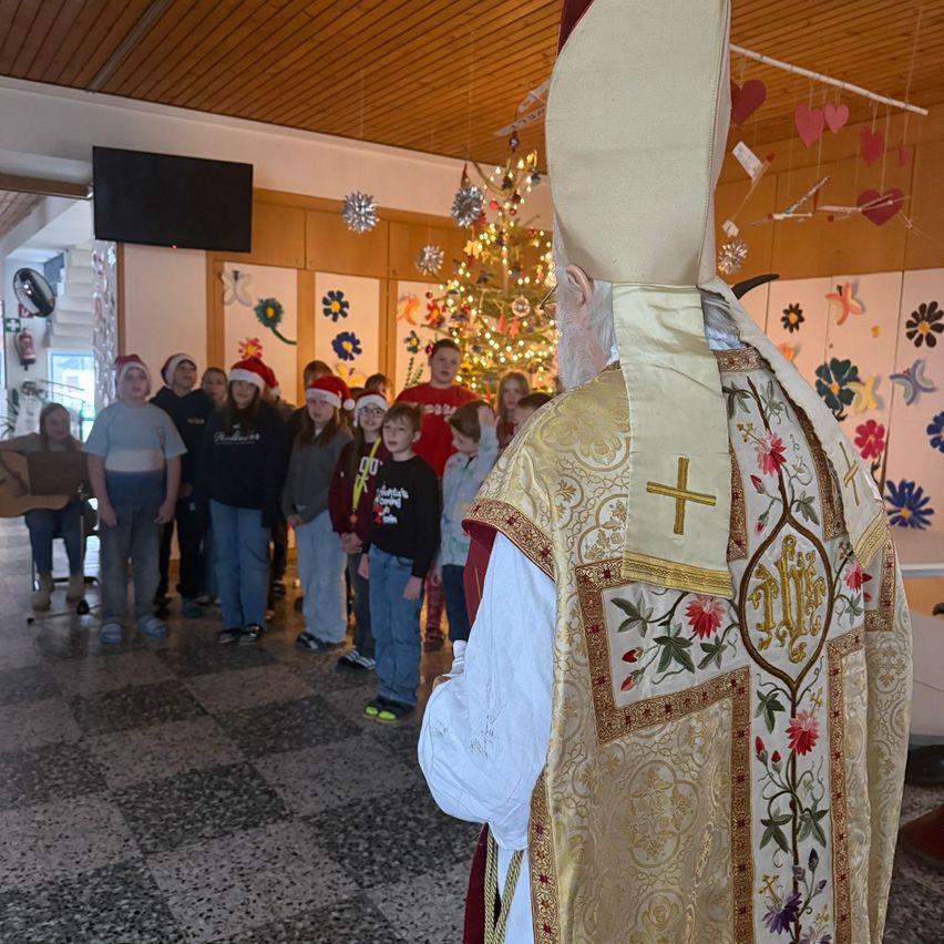 Ein Priester in prächtigen Gewändern steht vor einer Gruppe von Kindern, einige mit Weihnachtsmützen, in einem Raum mit einem Weihnachtsbaum und Dekorationen.