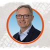 Ein lächelnder Mann mit Brille und Anzug in einem runden Rahmen. Er trägt ein weißes Hemd mit einem Logo auf der linken Brust.