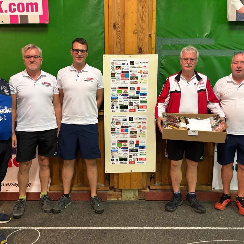 Fünf Männer stehen nebeneinander, wahrscheinlich bei einer Veranstaltung. Ein Mann hält eine Box mit Gegenständen. Sie tragen alle Turnschuhe. Der Mann in der Mitte trägt eine Brille und hat ein breites Lächeln im Gesicht. Hinter ihnen steht eine Wand mit einem Plakat.