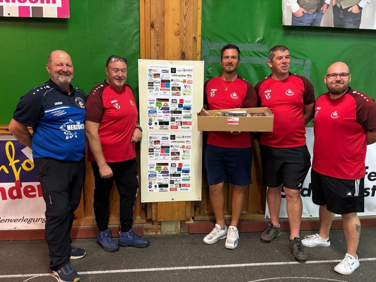 Fünf Männer in roten Shirts stehen vor einer grünen Wand mit einem Poster und lächeln für ein Foto. Der Mann in der Mitte hält eine Box.