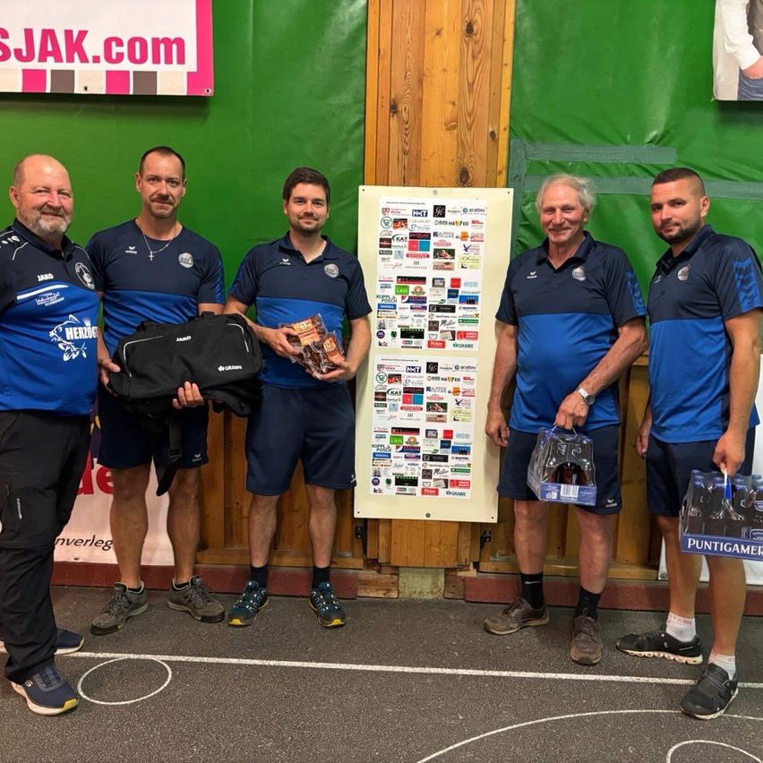 Fünf Männer stehen zusammen in einem Fitnessstudio und lächeln für die Kamera. Einer hält eine Tasche und eine Box mit Snacks, während ein anderer eine Getränkebox hält. Sie tragen identische blaue Uniformen. Hinter ihnen ist ein Plakat mit verschiedenen Logos.