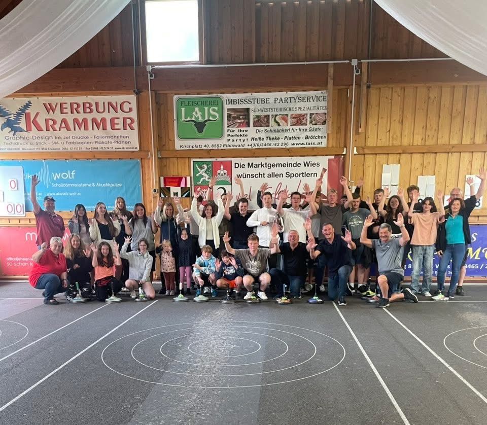 Eine Gruppe von Menschen in Sportkleidung posiert für ein Foto in einem Innenraum mit Bannern. Sie stehen auf einer Laufbahn und feiern möglicherweise ein Ereignis.