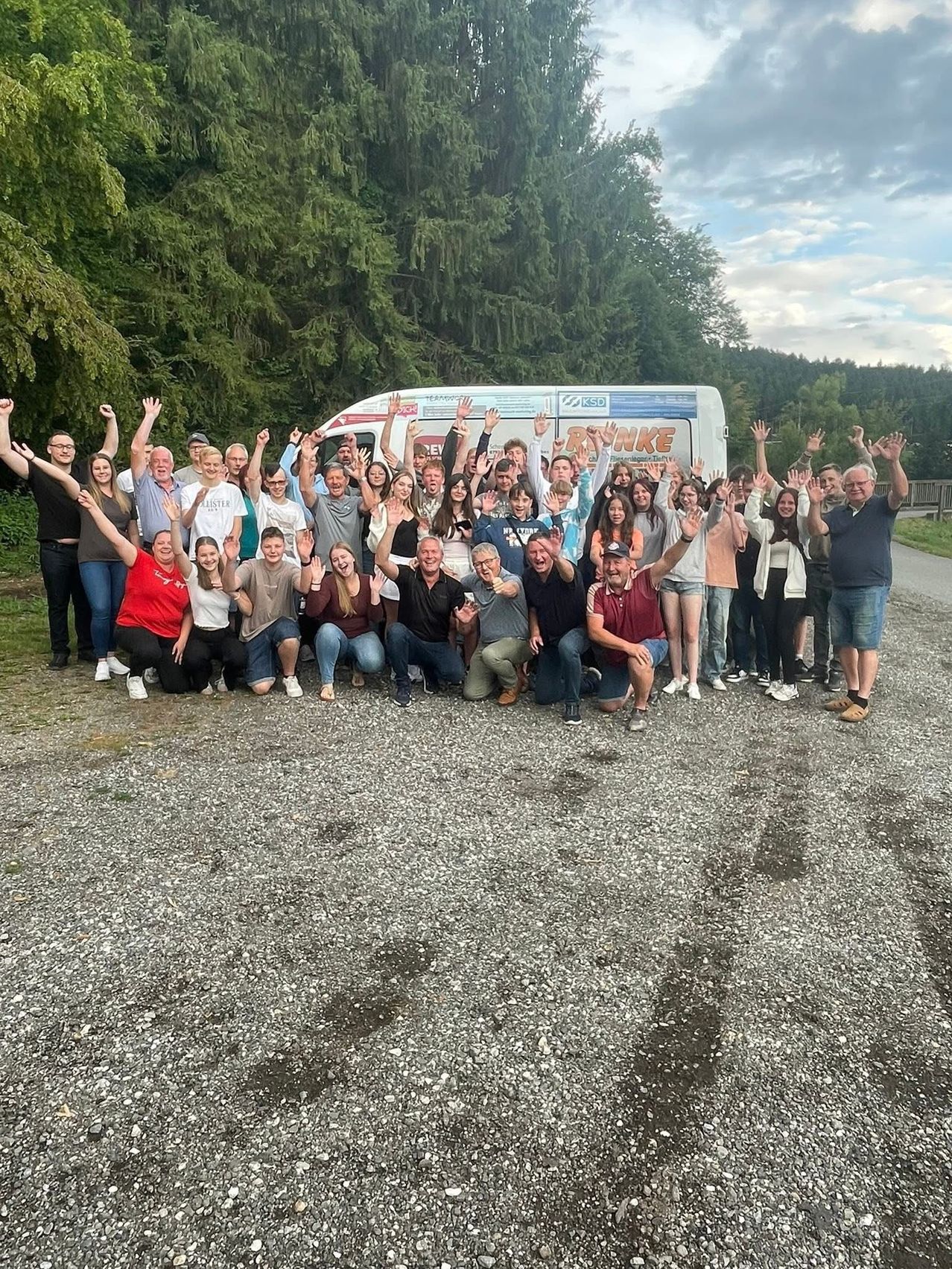 Eine Gruppe von Menschen, möglicherweise ein Team, posiert für ein Foto vor einem Van mit dem Wort 'RUNKE' darauf. Sie alle lächeln und heben ihre Hände.