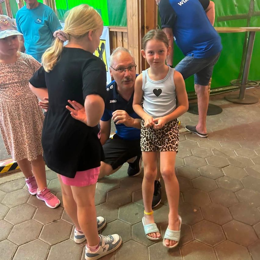 Eine Gruppe von Kindern und zwei Erwachsenen steht in einem gefliesten Raum, wobei ein Erwachsener kniet und mit einem jungen Mädchen in einem Herz-Tanktop spricht.