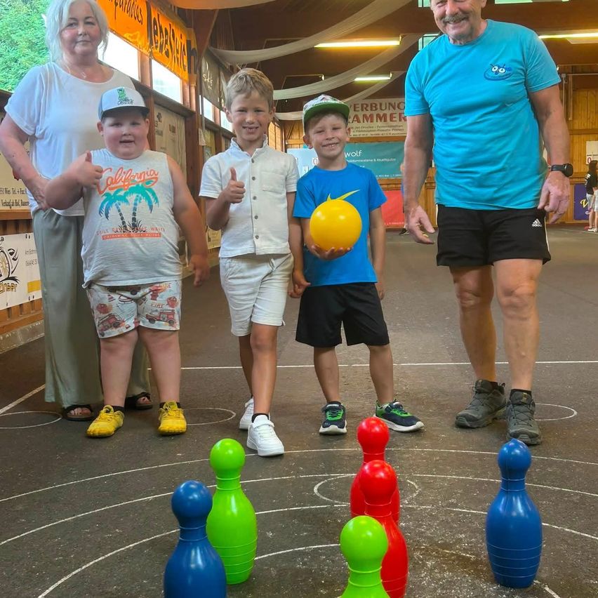 Eine vierköpfige Familie, bestehend aus zwei Erwachsenen und zwei Kindern, steht in einer Bowlingbahn mit bunten Bowlingpins auf dem Boden. Die Erwachsenen lächeln und die Kinder geben Daumen hoch.