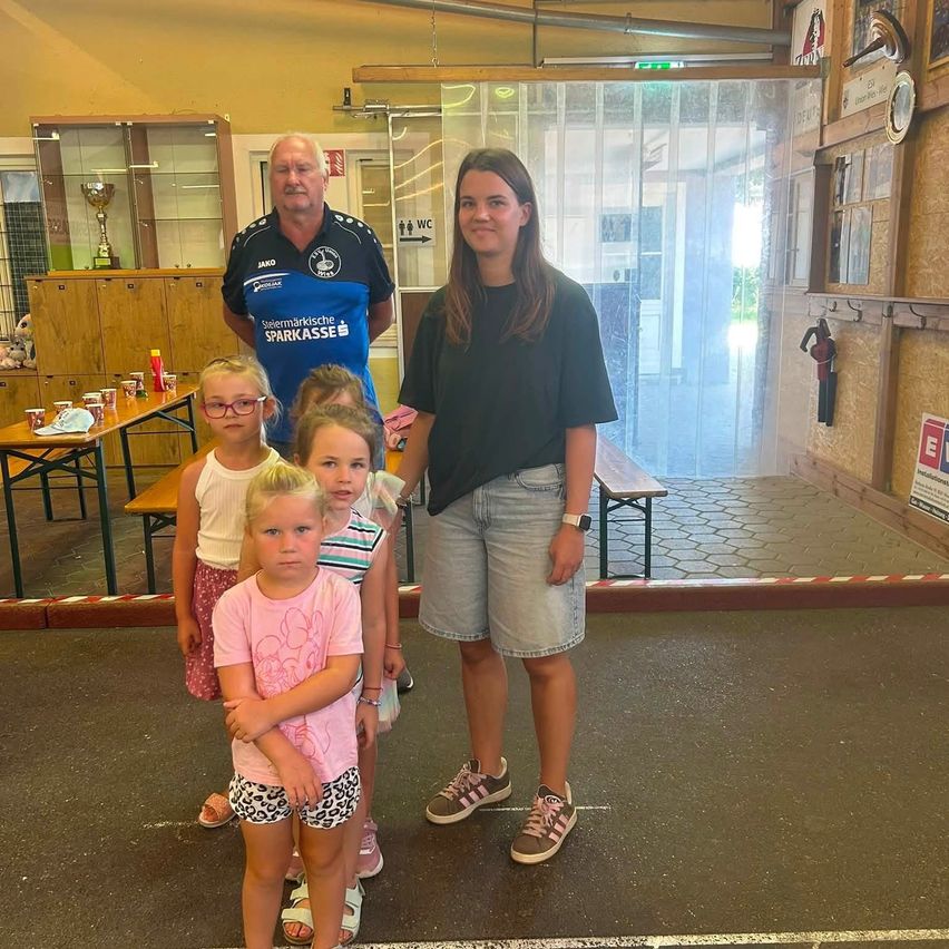 Eine Gruppe von Kindern und zwei Erwachsenen steht in einem Raum mit einem ausgestellten Pokal und einem Tisch mit Tassen. Die Frau trägt eine Uhr und Turnschuhe.